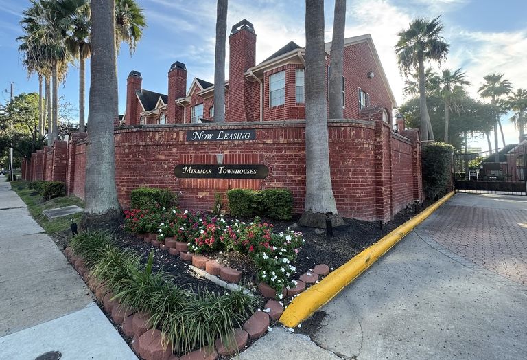 Red brick townhouses with "Now Leasing" sign, surrounded by palm trees and flowers, along a sunny sidewalk.