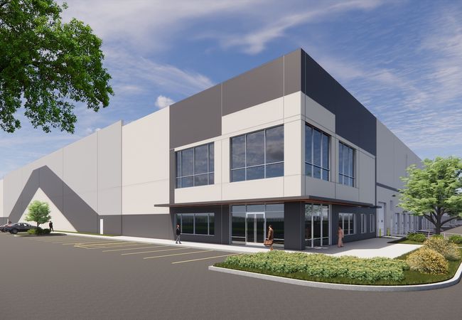 Modern industrial building with large windows, surrounded by a paved parking area and landscaped greenery under a partly cloudy sky.