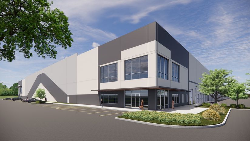Modern industrial building with large windows, surrounded by a paved parking area and landscaped greenery under a partly cloudy sky.