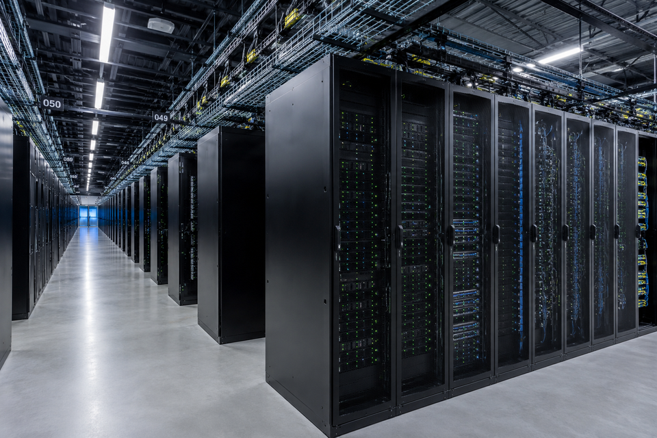 A spacious, modern server room with rows of black server racks and overhead cable trays under bright fluorescent lighting.