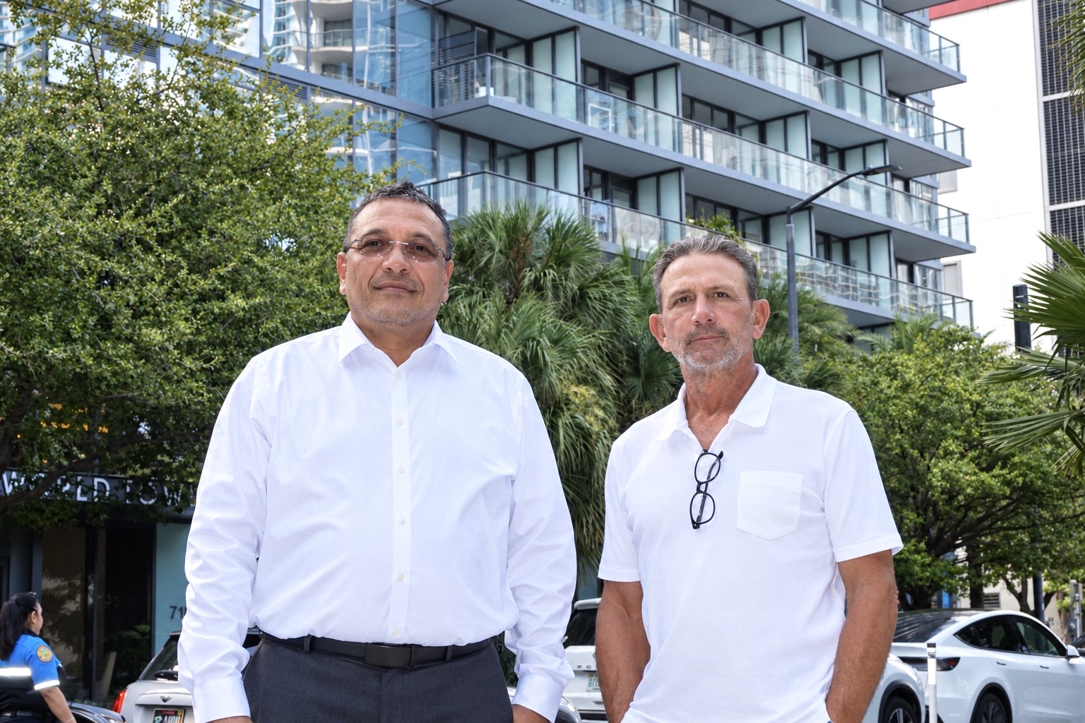 Two men stand outdoors in front of a modern building with glass balconies and trees in the background, wearing a white shirt and a white polo shirt.