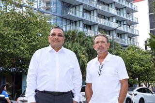 Two men stand outdoors in front of a modern building with glass balconies and trees in the background, wearing a white shirt and a white polo shirt.