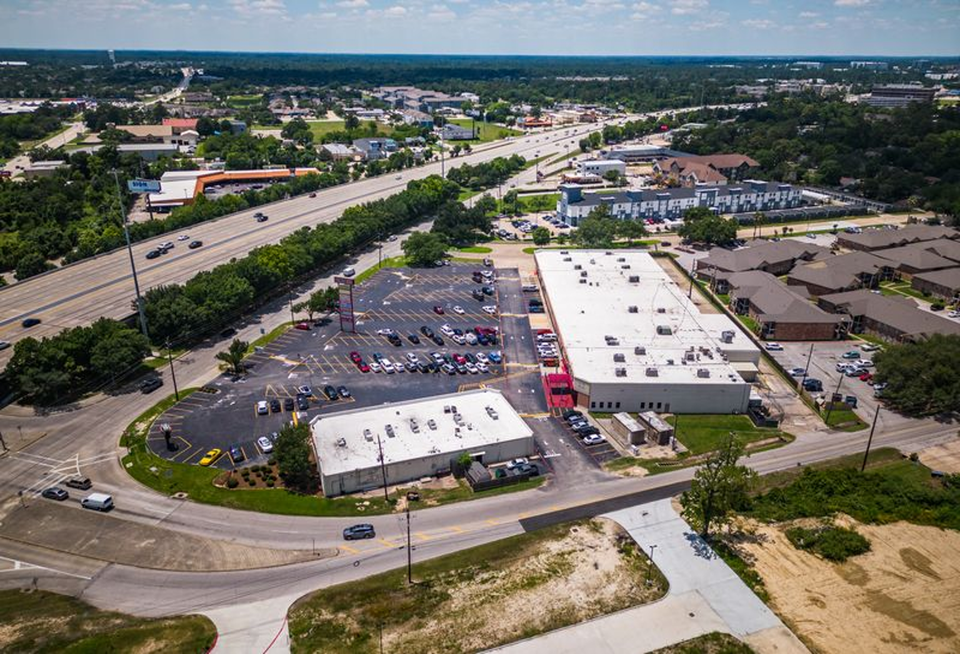 An aerial view of a commercial area with parking lots, roads, residential buildings, and surrounding greenery under a partly cloudy sky.