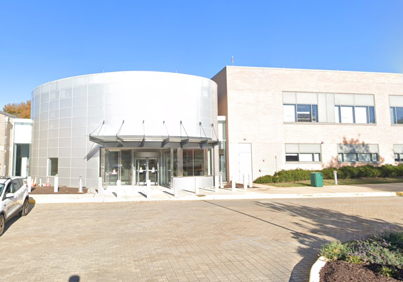 Modern building with a cylindrical glass and metal entrance, flat-roofed wing on the right, and a paved area in front. Clear sky overhead.
