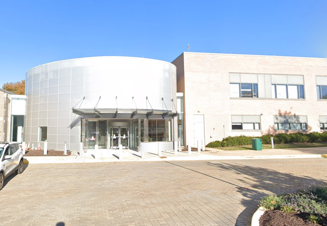 Modern building with a cylindrical glass and metal entrance, flat-roofed wing on the right, and a paved area in front. Clear sky overhead.