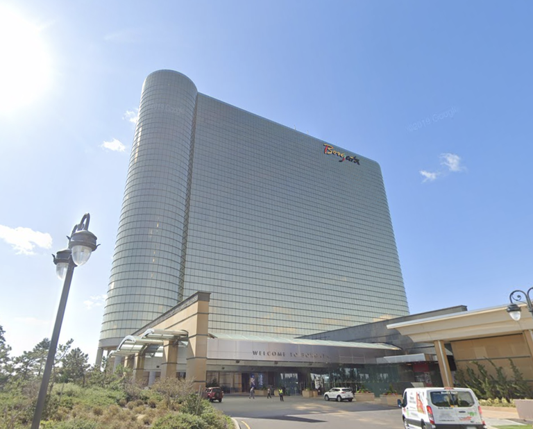 Tall glass building with "Borgata" sign, blue sky, sunlight, and a driveway with cars and lampposts in the foreground.