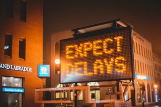 An illuminated road sign reads "EXPECT DELAYS" at night, set against a backdrop of urban buildings.