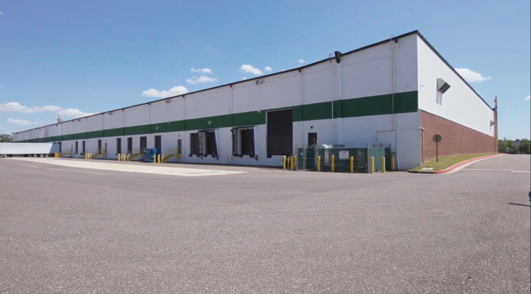 Large white warehouse with green stripe, multiple loading bays, and a trailer parked alongside. Blue sky and few clouds above.