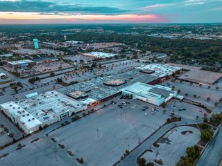 Aerial view of a sprawling shopping mall complex surrounded by large, mostly empty parking lots at dusk, under a cloudy sky with cityscape in the background.