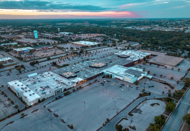 Aerial view of a sprawling shopping mall complex surrounded by large, mostly empty parking lots at dusk, under a cloudy sky with cityscape in the background.
