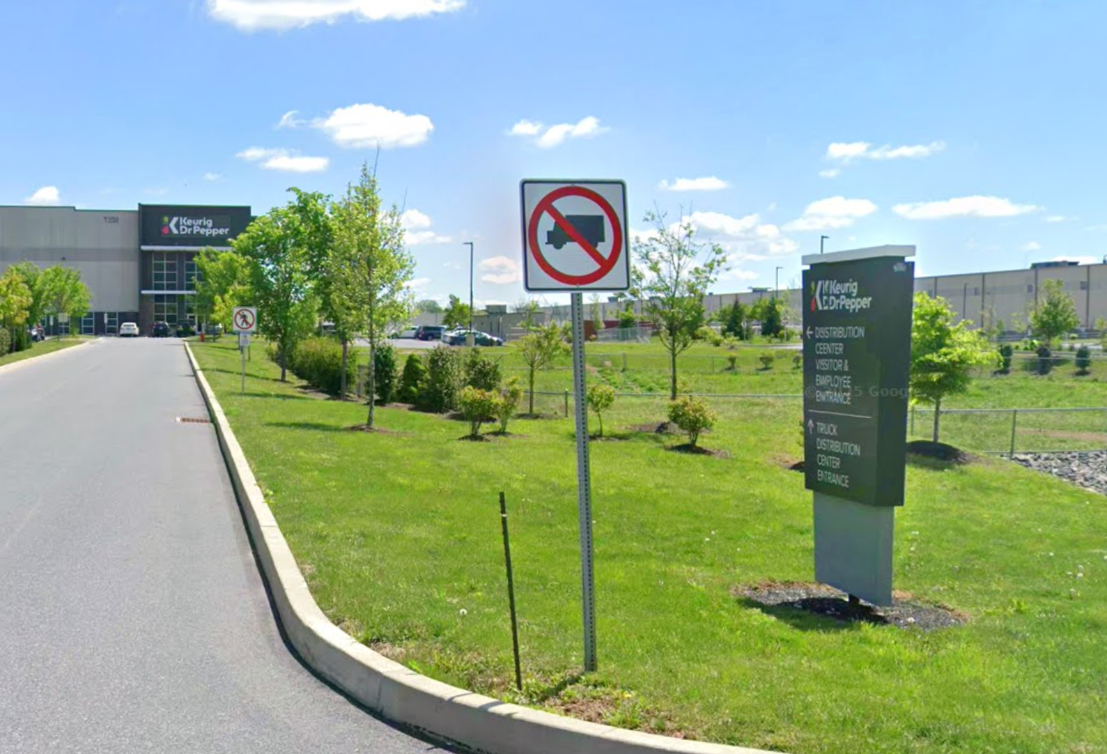 Driveway leading to a distribution center with signs prohibiting trucks and guiding to entrances, surrounded by green lawns.