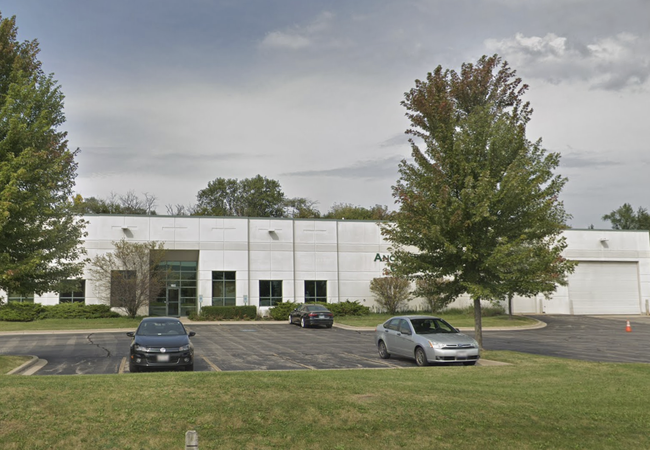 White industrial building with multiple cars parked in front, surrounded by trees.