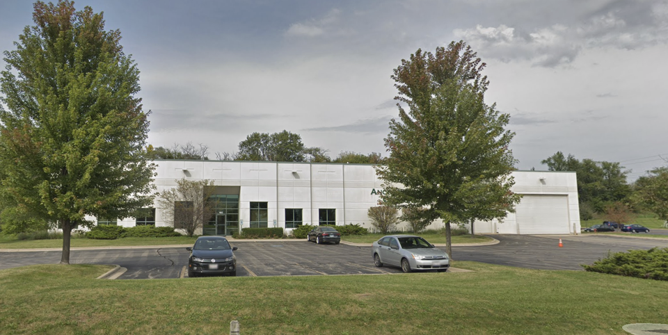 White industrial building with multiple cars parked in front, surrounded by trees.