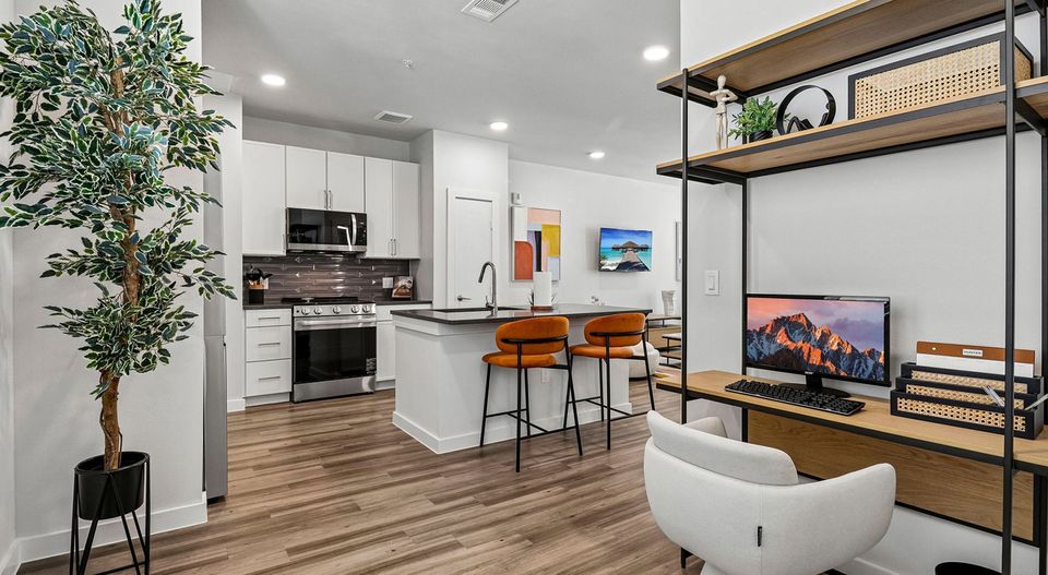 Modern open-plan kitchen and workspace with white cabinets, orange bar stools, wood floor, decorative plant, and wall art.