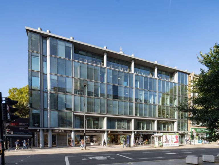 A modern multi-story glass building with shops at street level, situated on a bustling city street under a clear blue sky.