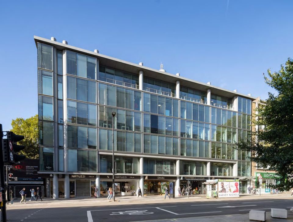 A modern multi-story glass building with shops at street level, situated on a bustling city street under a clear blue sky.