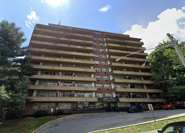 A multi-story brick apartment building with balconies under a bright blue sky, cars parked in front, and surrounded by trees.