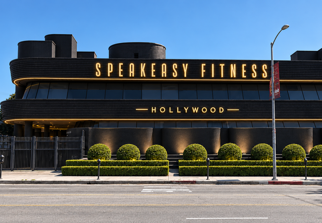 Modern black building labeled "Speakeasy Fitness Hollywood" with landscaping and a street in the foreground.