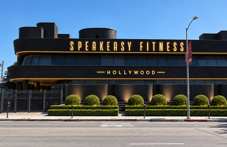 Modern black building labeled "Speakeasy Fitness Hollywood" with landscaping and a street in the foreground.