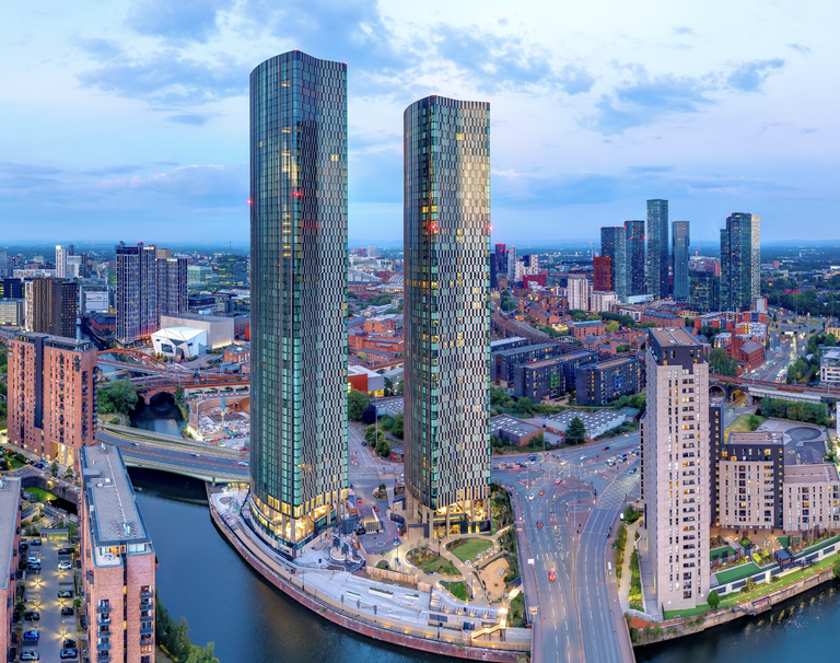 Two modern skyscrapers tower over an urban landscape by a river, surrounded by roads and smaller buildings under a cloudy sky.