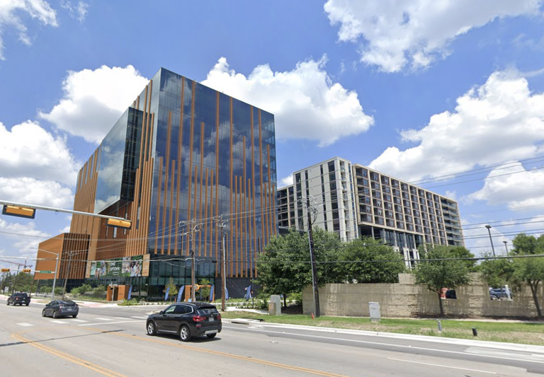 A modern glass building with orange vertical accents next to a residential block, set against a blue sky with clouds. Cars drive in the foreground.