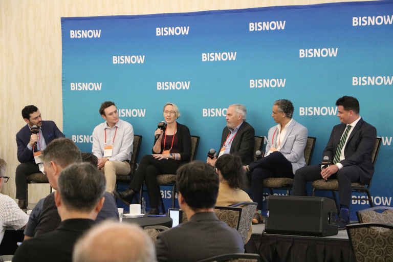 A panel of six people sits on stage in front of a blue "BISNOW" backdrop, engaged in a discussion with microphones.