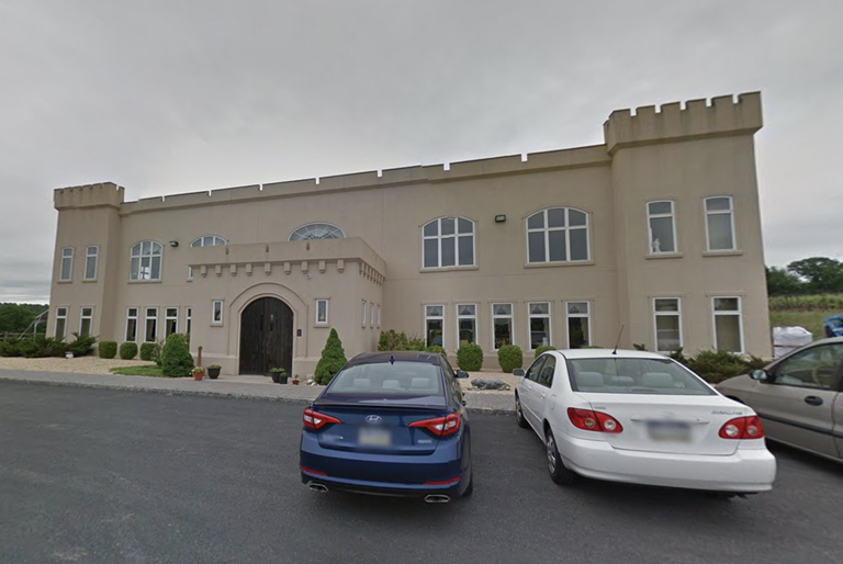 Beige building with castle-like features, large arched entrance, and multiple windows. Three cars are parked in front on an overcast day.
