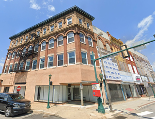 Historic multi-story brick building with arched windows, fire escape, and adjoining shops, situated on a city street with clear blue sky above.