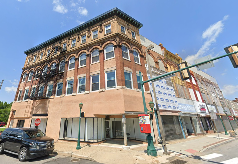 Historic multi-story brick building with arched windows, fire escape, and adjoining shops, situated on a city street with clear blue sky above.