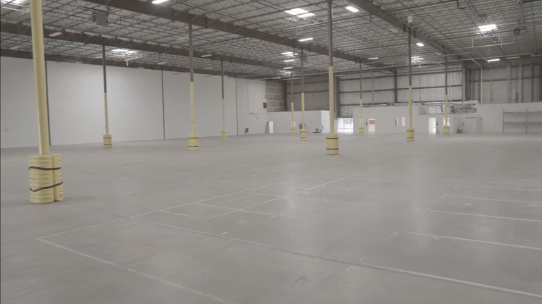 Spacious empty warehouse interior with concrete floor, high ceiling, steel beams, and yellow safety-wrapped pillars.