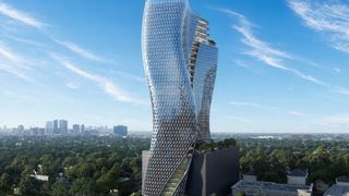 A modern, twisted skyscraper with a glass facade stands against a blue sky, surrounded by greenery and urban buildings in the distance.