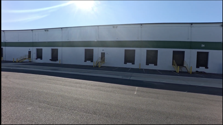 A large warehouse with a white exterior and green stripe, featuring loading docks and entrance stairs under bright sunlight.