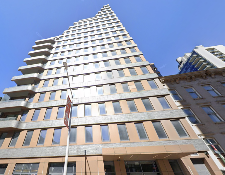 Tall modern building with a unique stepped design, reflective windows, and light tan facade against a clear blue sky.