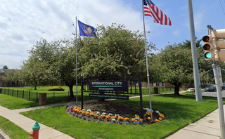 Two flags next to the International City Apartments sign on a grassy corner with flowerbeds, flanked by a stoplight and sidewalk under a partly cloudy sky.