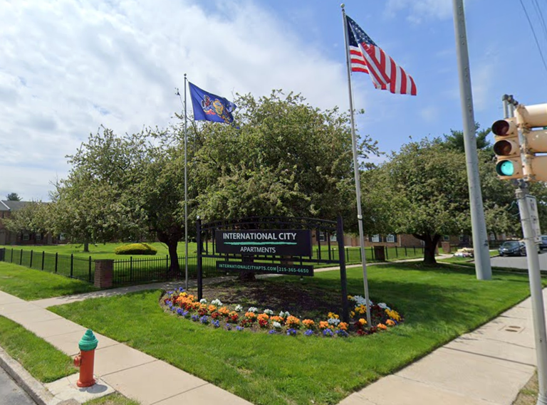 Two flags next to the International City Apartments sign on a grassy corner with flowerbeds, flanked by a stoplight and sidewalk under a partly cloudy sky.