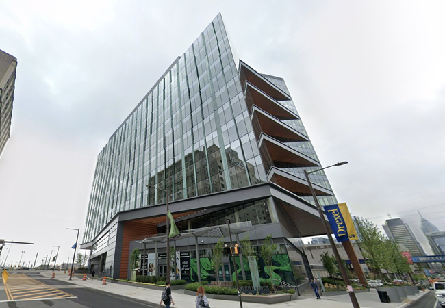 Modern glass building with angled architectural design, located on a city street with pedestrians and a Drexel banner visible.