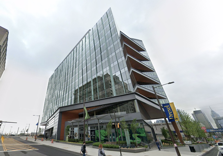 Modern glass building with angled architectural design, located on a city street with pedestrians and a Drexel banner visible.