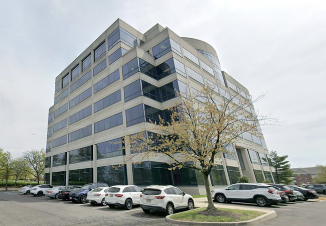 Office building with large windows, surrounded by parked cars and trees. The sky is overcast.