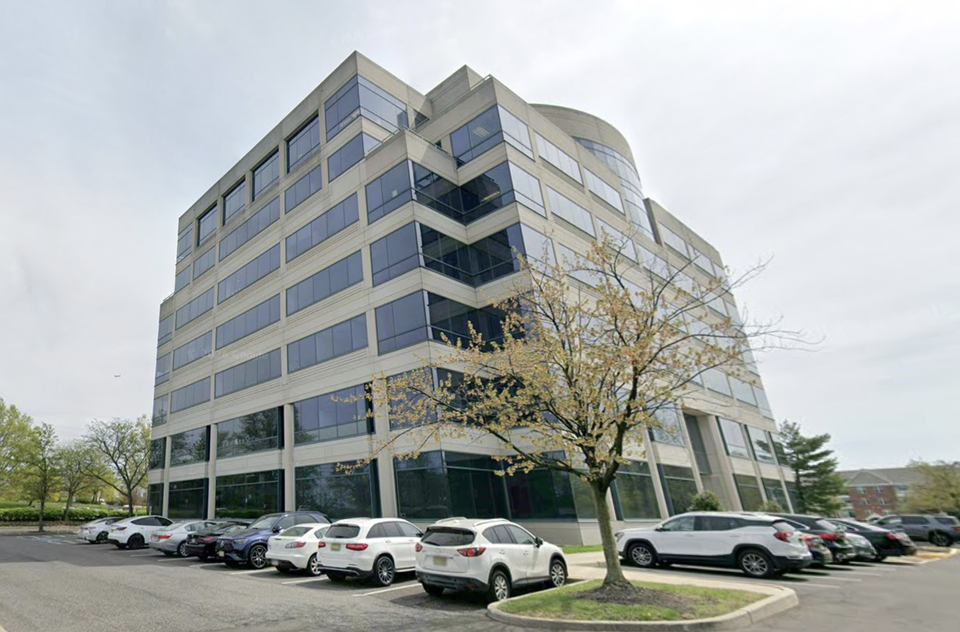 Office building with large windows, surrounded by parked cars and trees. The sky is overcast.