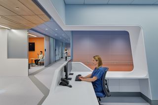 Woman in scrubs sits at a modern reception desk with computers. Two people are visible in an adjacent room with orange and blue walls.