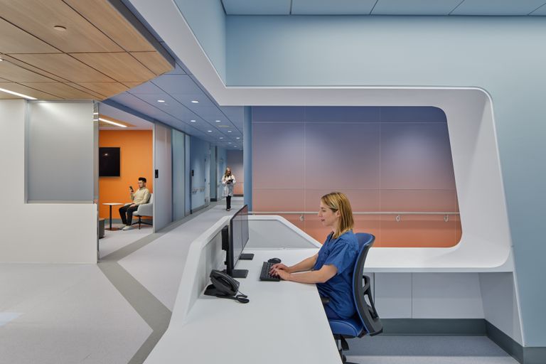Woman in scrubs sits at a modern reception desk with computers. Two people are visible in an adjacent room with orange and blue walls.