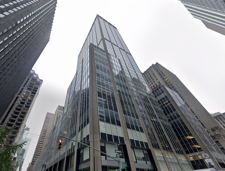 Tall modern skyscraper with glass facades surrounded by other buildings under a cloudy sky. A traffic light is visible in the foreground.