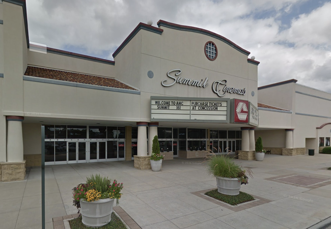 Entrance of Summit Cinemas, featuring marquee signs with movie information and a ticket purchase notice, set against a cloudy sky. Large flower pots in front.
