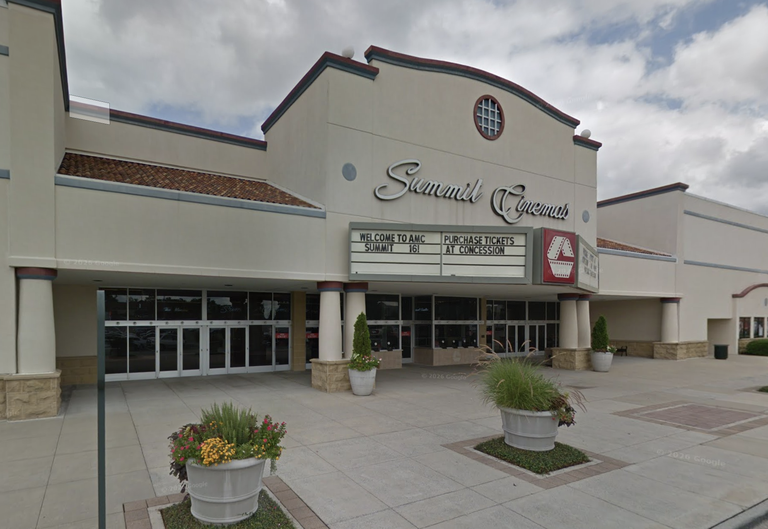 Entrance of Summit Cinemas, featuring marquee signs with movie information and a ticket purchase notice, set against a cloudy sky. Large flower pots in front.