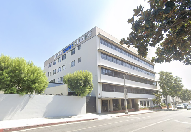 A four-story beige building labeled "Sunset Gower Studios" with large windows, surrounded by trees and situated on a sunny street.