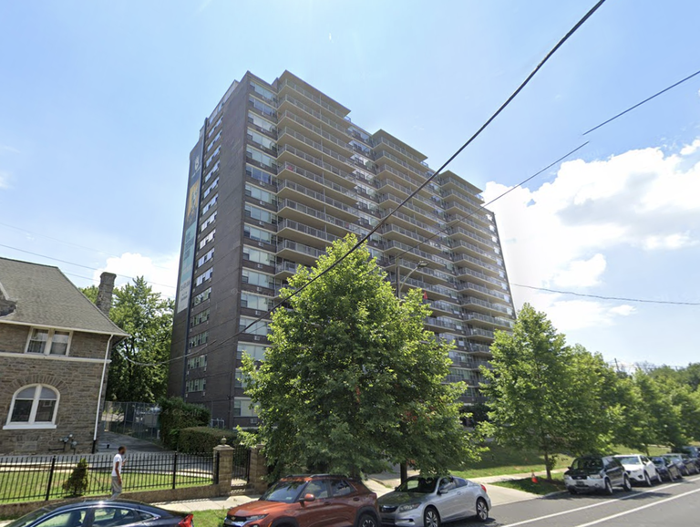 A tall apartment building with multiple balconies stands next to a large tree and a smaller stone house, with parked cars lining the street below.