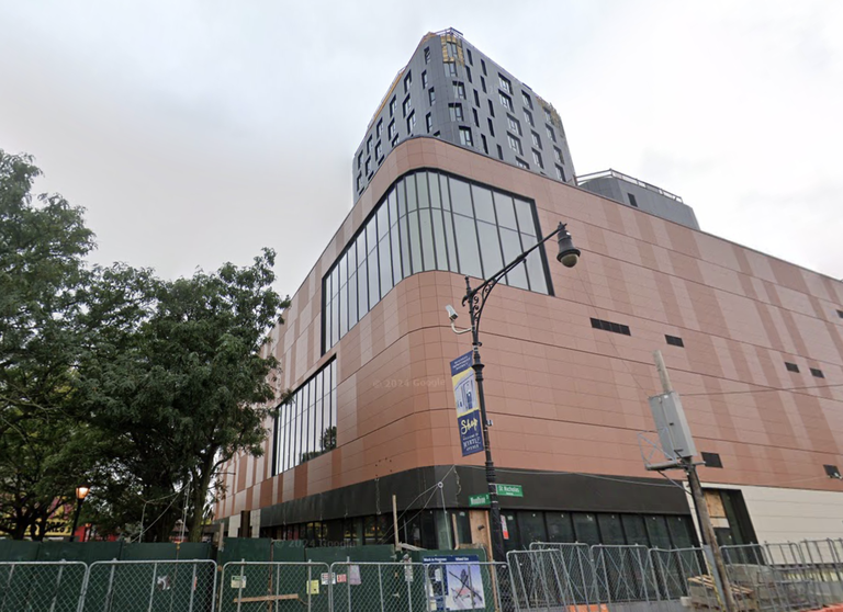 Modern multi-story building with large windows, curved facade, and a fenced construction area at street level, next to a tree and streetlight.