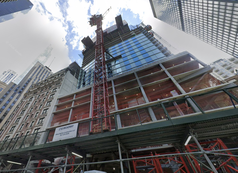A high-rise building under construction with a prominent red crane, surrounded by other tall buildings and a cloudy sky above.