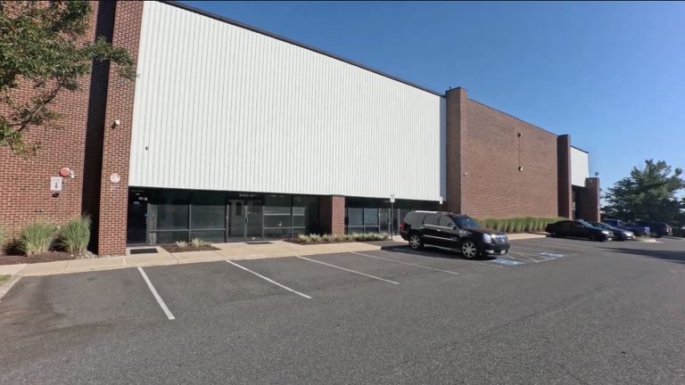 A large brick and white industrial building with a mostly empty parking lot and a few parked vehicles under a clear blue sky.