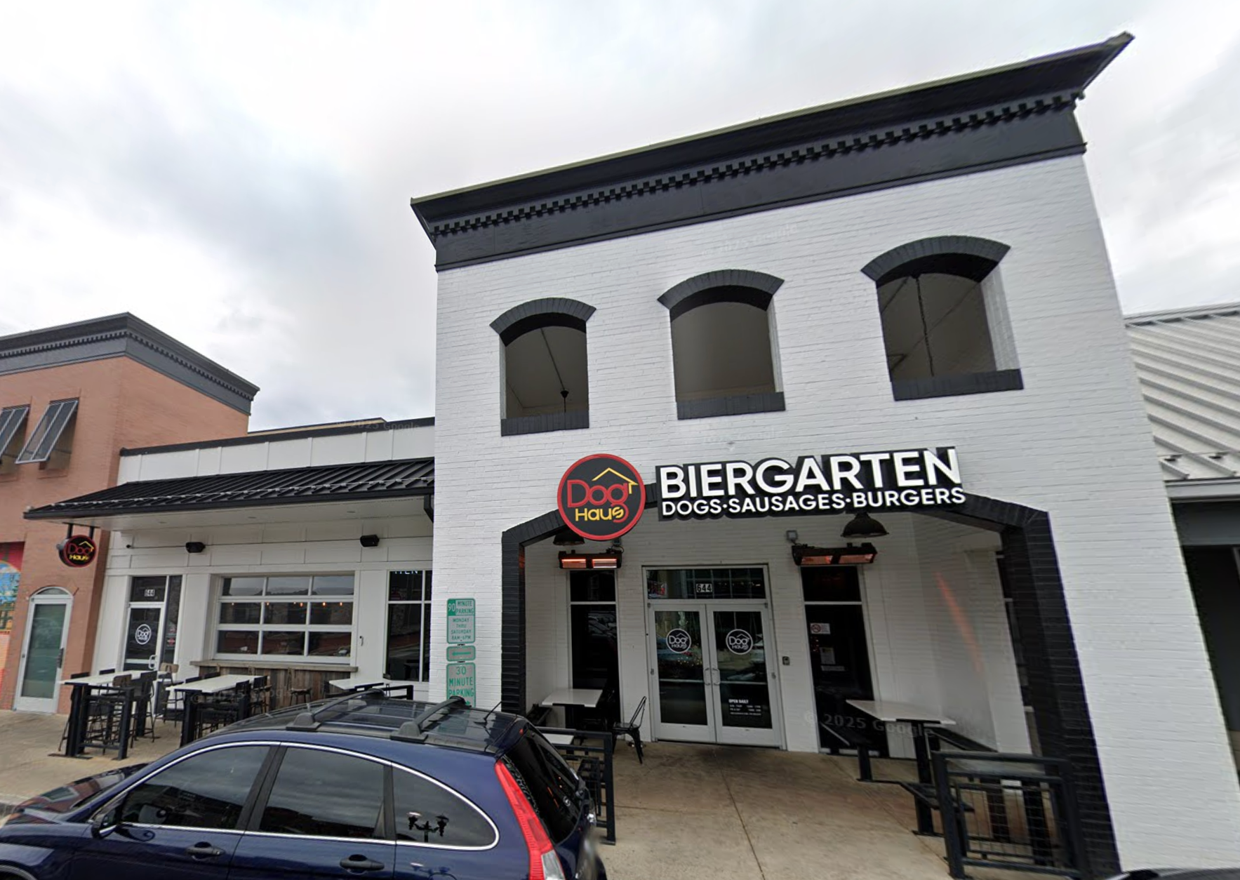 Exterior of a building with a sign reading "Dog Haus Biergarten Dogs, Sausages, Burgers." Outdoor seating and a parked car in front.